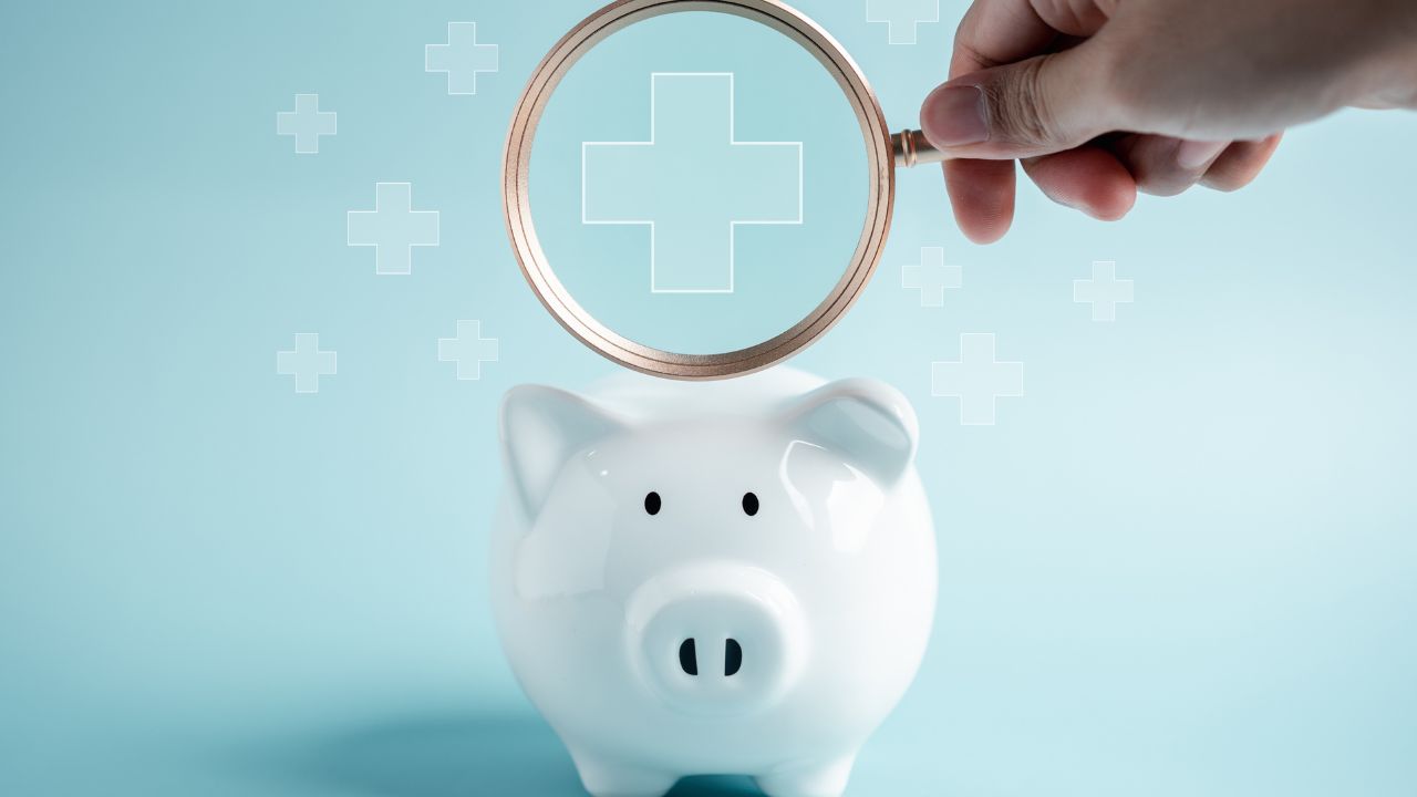 Person holding a magnifying glass over a white piggy bank with medical crosses above it